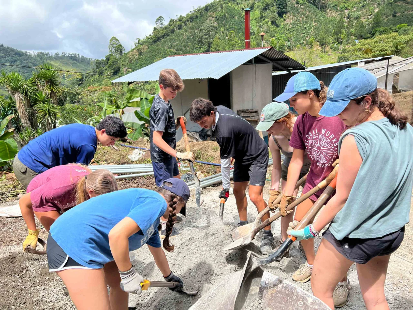 A group of people are using shovels to mix and move sand or gravel at a construction site in a rural, hilly area. They appear to be working together on a project outside.