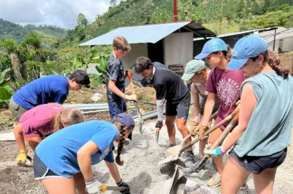 A group of people are using shovels to mix and move sand or gravel at a construction site in a rural, hilly area. They appear to be working together on a project outside.