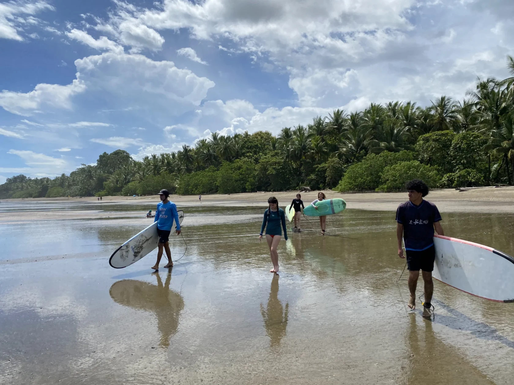 Four people in a Teen Travel Program walk along a sandy beach carrying surfboards, with palm trees and a cloudy blue sky in the background.