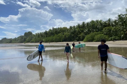 Four people in a Teen Travel Program walk along a sandy beach carrying surfboards, with palm trees and a cloudy blue sky in the background.