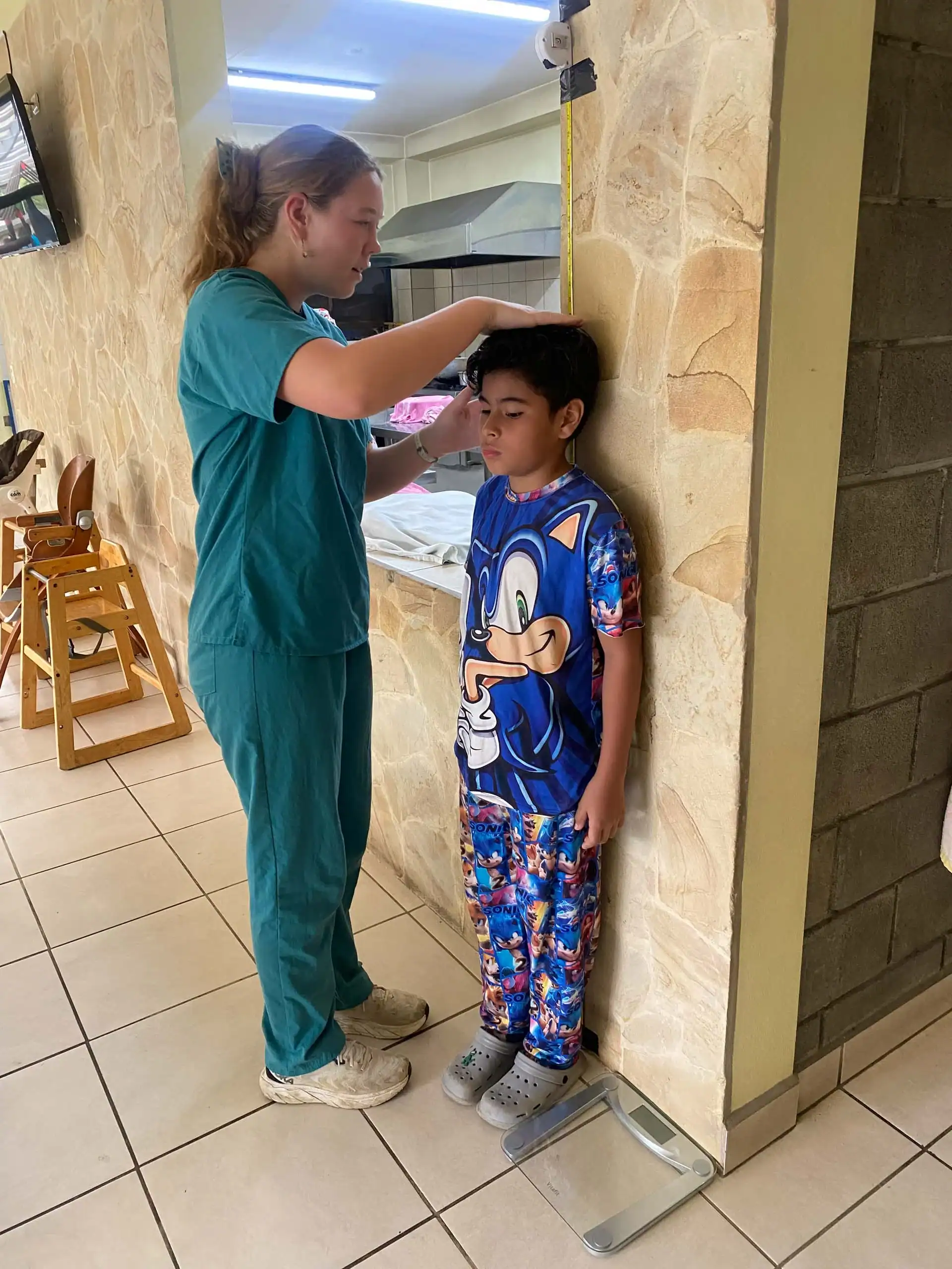 A healthcare worker, part of a pre med travel program, measures the height of a boy in Sonic pajamas against a wall using a stadiometer in a tiled room.
