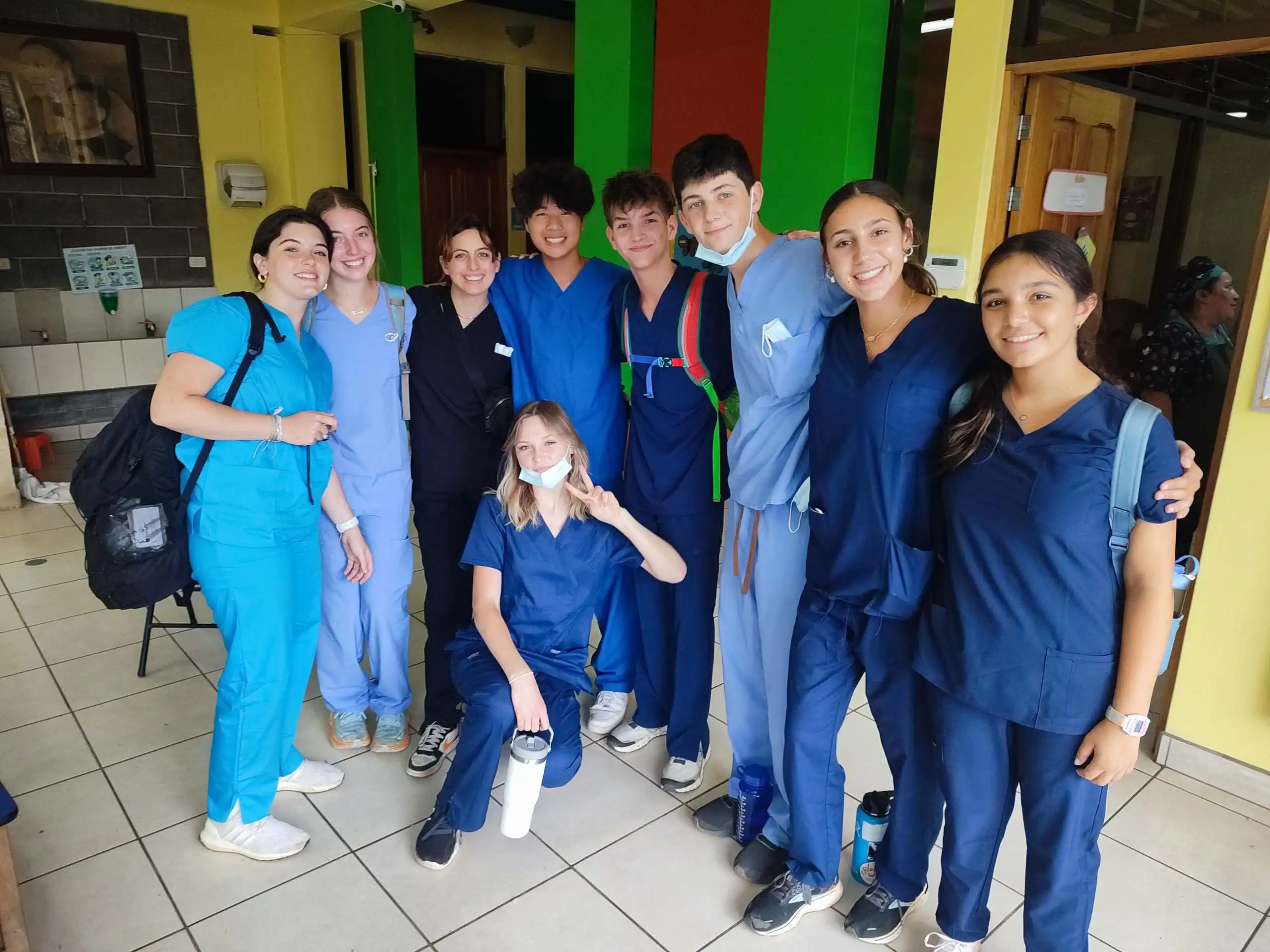 A group of nine young people in scrubs, part of a pre med travel program, pose together indoors, smiling at the camera. Some hold backpacks and water bottles, while one person flashes a peace sign.