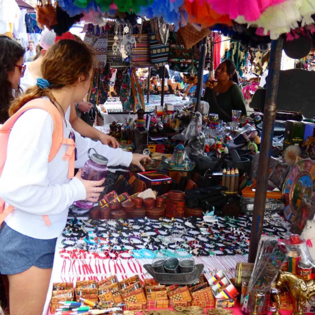 People browsing a colorful market stall with various handmade crafts, jewelry, and souvenirs on display.
