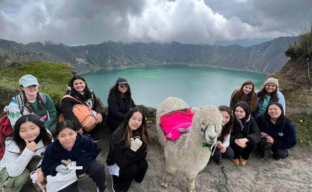 A group of people kneels on a path near a llama with a pink saddle, in front of a lake surrounded by mountains under a cloudy sky.