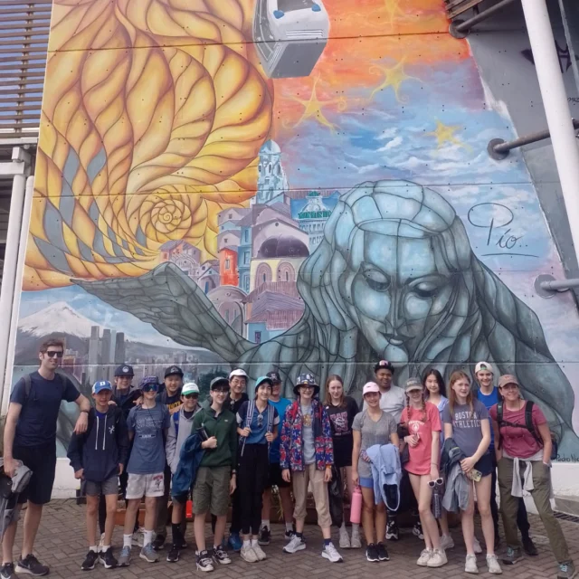 A group of people pose in front of a colorful mural featuring a large figure and buildings with a blend of abstract and realistic elements.