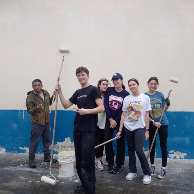 A group of six people holding paint rollers stand in front of a freshly painted wall, with a blue section at the bottom. They are posing for the photo and smiling.
