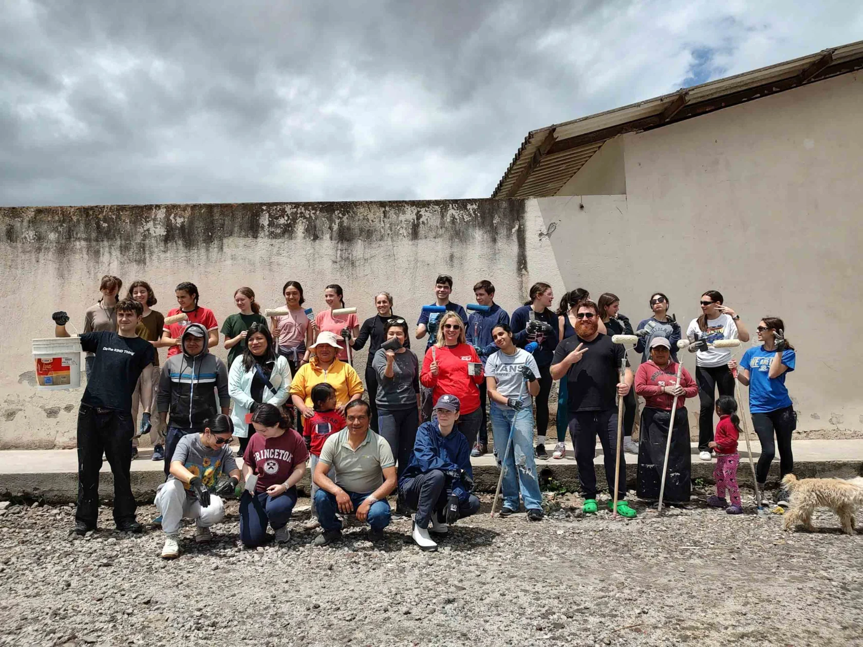 A group of people pose for a photo outdoors against a weathered wall. Some hold tools and a dog is present.