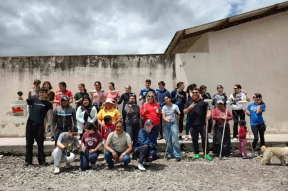 A group of people pose for a photo outdoors against a weathered wall. Some hold tools and a dog is present.