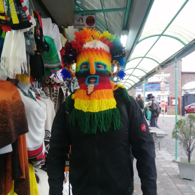 Person wearing a colorful, fringed mask in a marketplace, surrounded by hanging textiles.