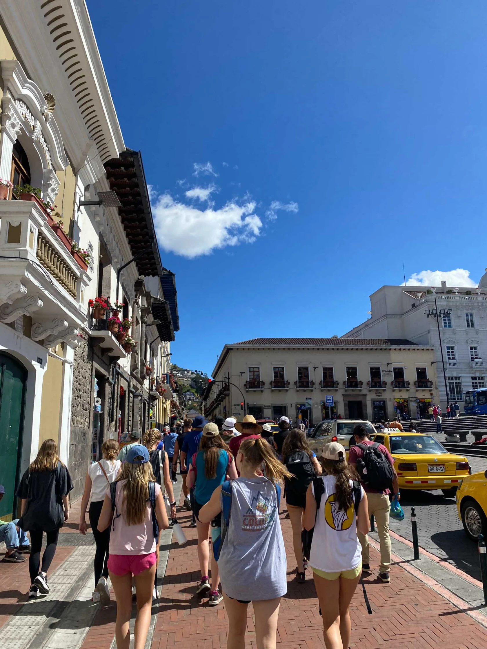 A group of people walk down a sunny street with historic buildings, taxis, and a blue sky.