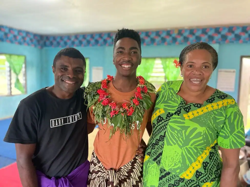 Three people stand indoors, smiling at the camera. The person in the center wears a floral garland, while the others wear brightly patterned clothing—capturing a joyful moment during a student travel program by Global Works.
