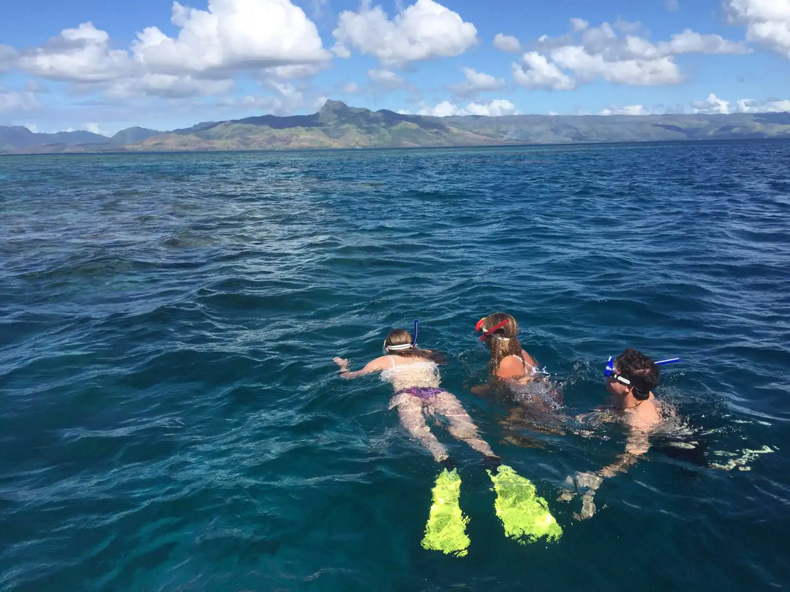 Three people snorkeling in clear blue water with a mountainous island landscape in the background.