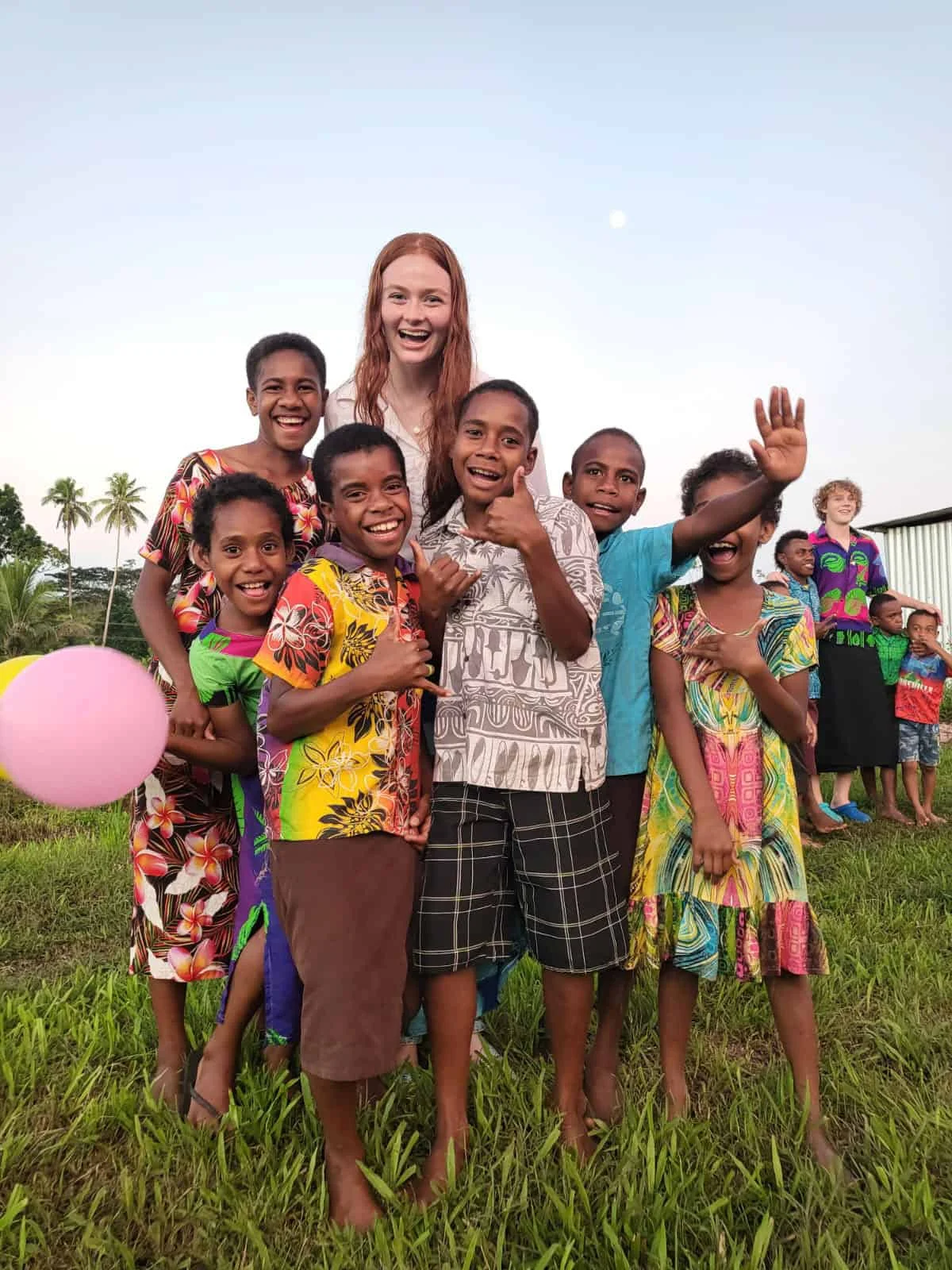A group of smiling children and an adult stand together on grass, some giving thumbs up, with a pink balloon and trees in the background—capturing joy during a student travel program by Global Works.