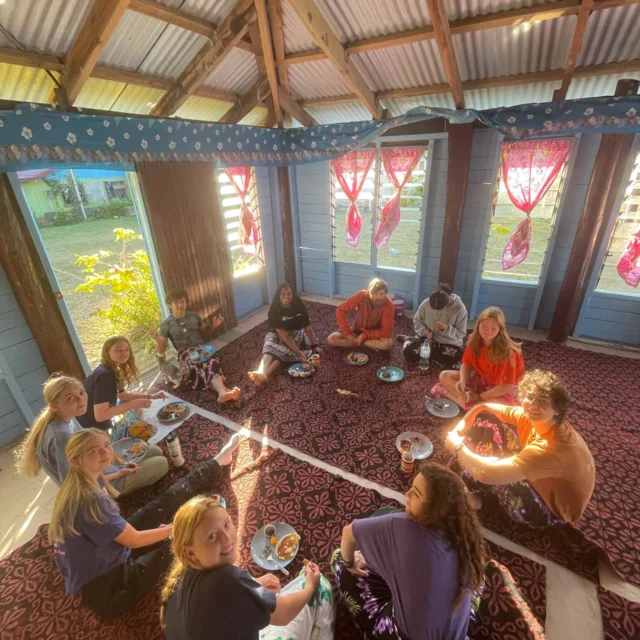 A group of people sit on a decorated floor in a room with red curtains, sharing a meal from plates placed in front of them.