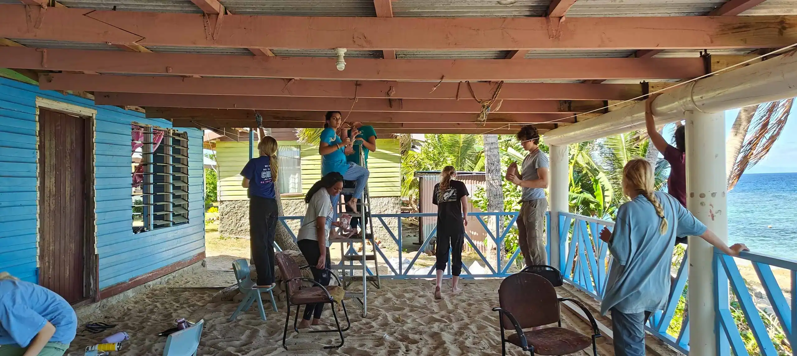 People renovating a covered patio by the sea, working on the roof and railings. Chairs and tools are scattered around.