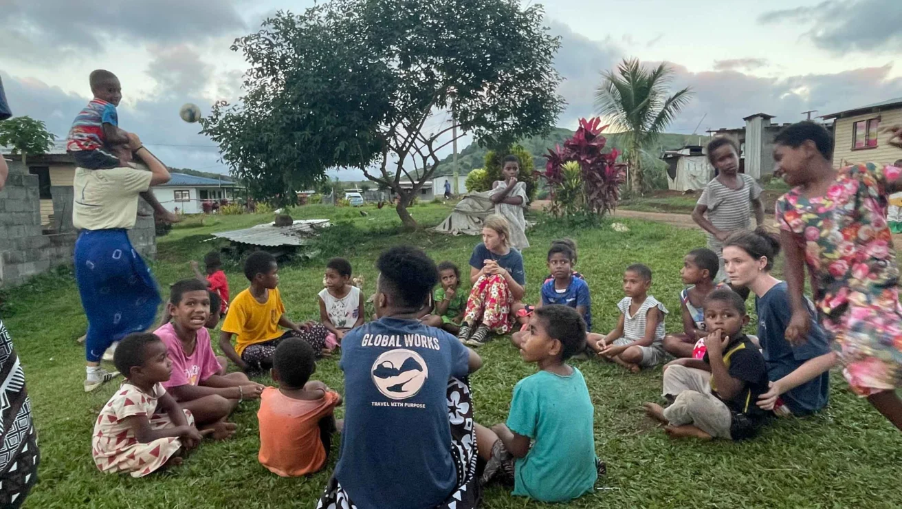 A group of children and adults sit and stand in a grassy area, with trees and buildings in the background.