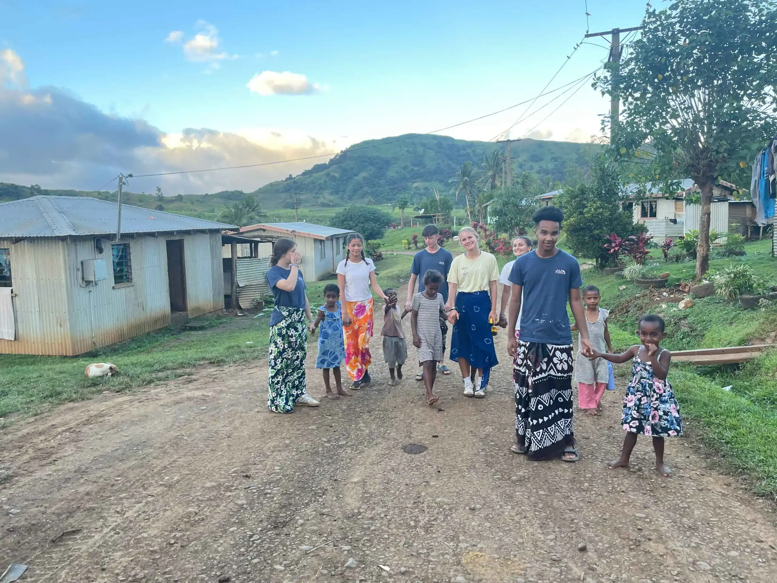A group of adults and children walk together on a dirt road in a rural village with houses, greenery, and hills in the background, capturing the spirit of a student trip to Fiji.