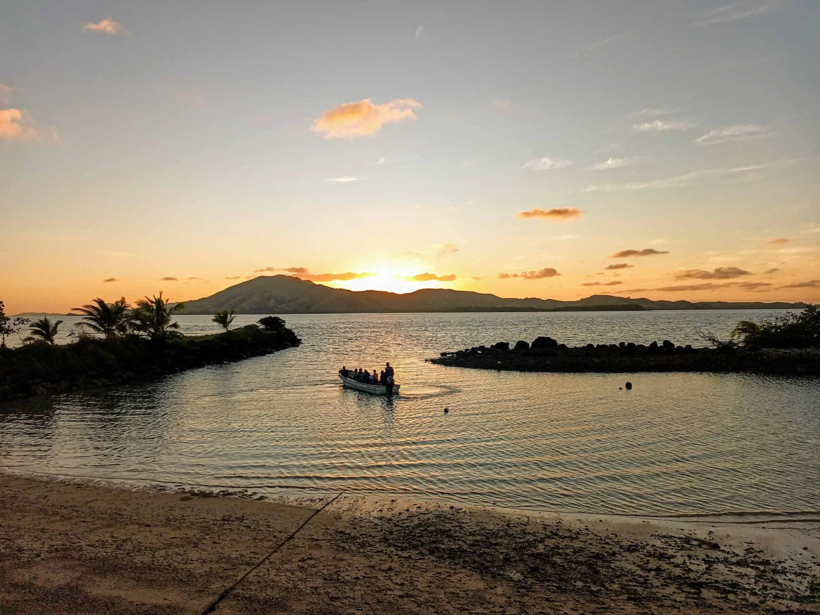 A small boat with people on calm water during a sunset, surrounded by palm trees and distant mountains.