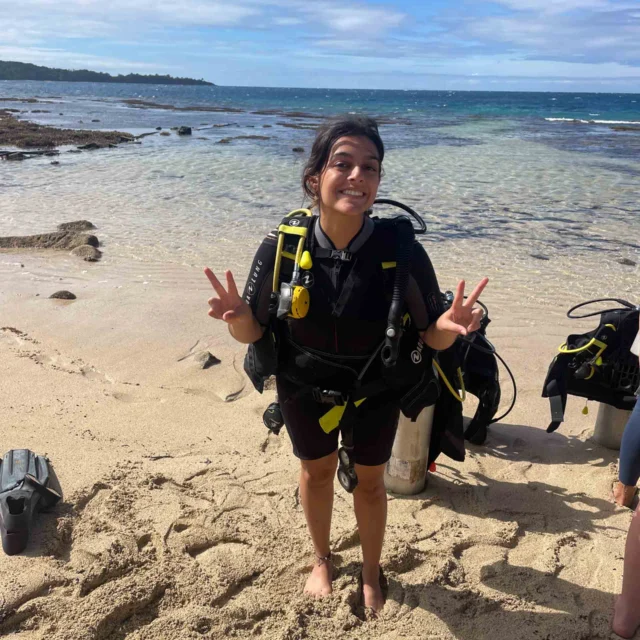 A person in a wetsuit and scuba gear stands on a sandy beach, smiling and making peace signs. Ocean waves are visible in the background.