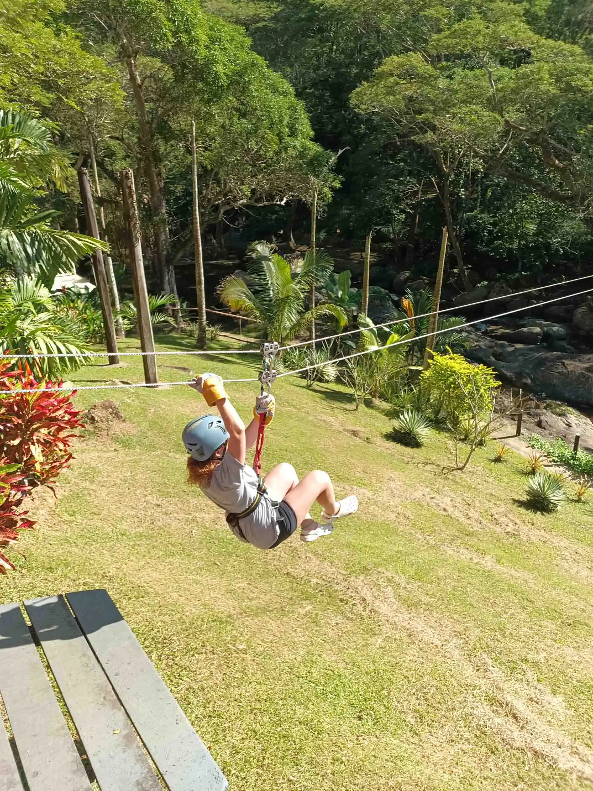 A person wearing a helmet and gloves rides a zipline over a grassy area surrounded by trees and plants in daylight, capturing the thrill of adventure on a Fiji student trip.