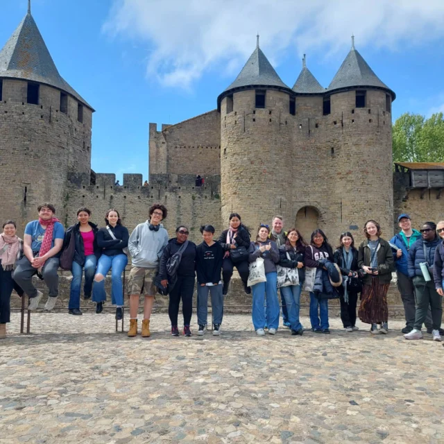 A group of people pose in front of a historic stone castle with round towers and conical roofs on a cobblestone plaza. The sky is partly cloudy.