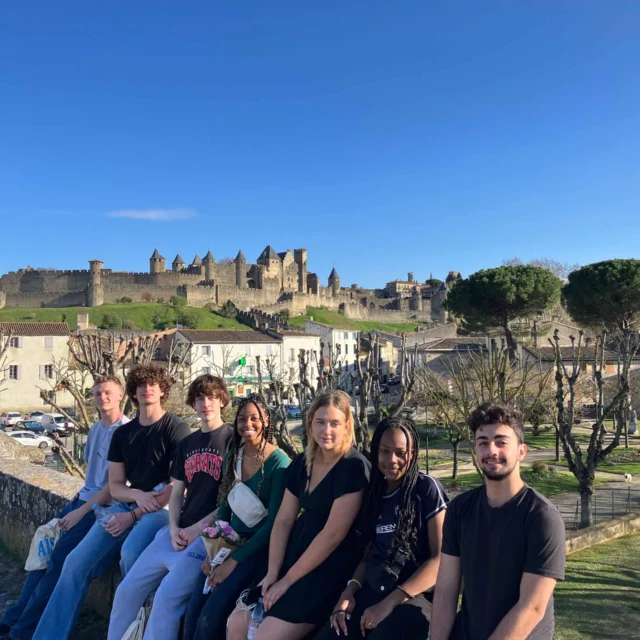 A group of people sitting on a stone ledge with a historic castle in the background under a clear blue sky.