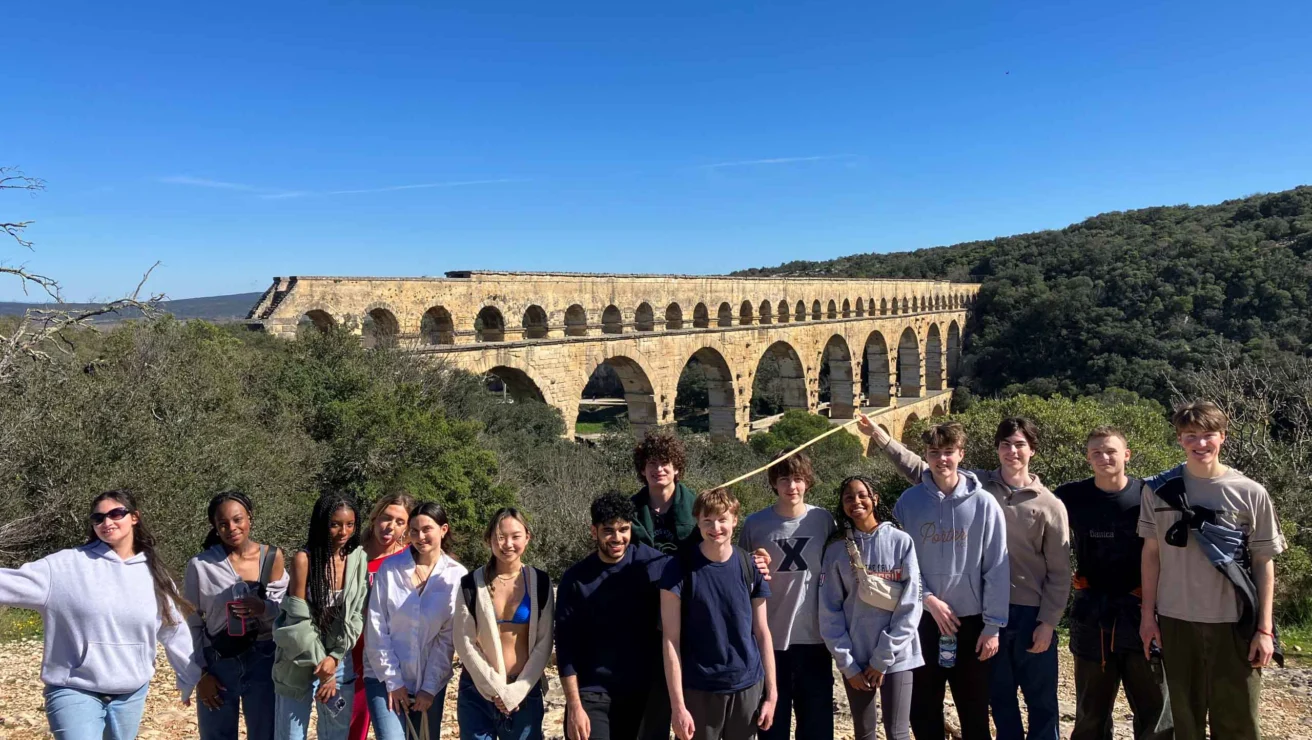 A group of people stands in front of the Pont du Gard aqueduct on a sunny day, with trees and blue sky in the background.