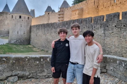 Three young men standing arm in arm on a stone walkway, with an ancient castle featuring round towers and high walls in the background.