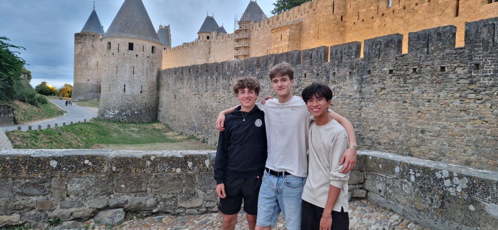 Three young men standing arm in arm on a stone walkway, with an ancient castle featuring round towers and high walls in the background.