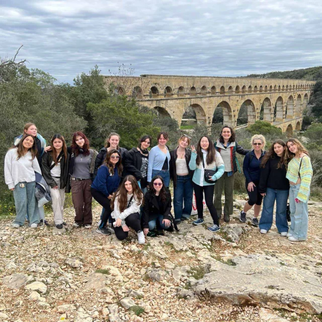 A group of people posing in front of an ancient stone aqueduct surrounded by greenery and a cloudy sky.