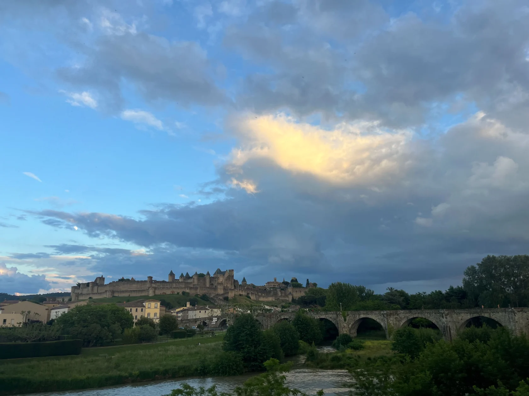 A scenic view of Carcassonne with a medieval fortress, houses, lush greenery, a river, and a stone bridge under a partly cloudy sky.