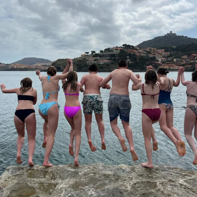 A group of people in swimwear are jumping into the water from a rocky ledge, with hills and buildings visible in the background.