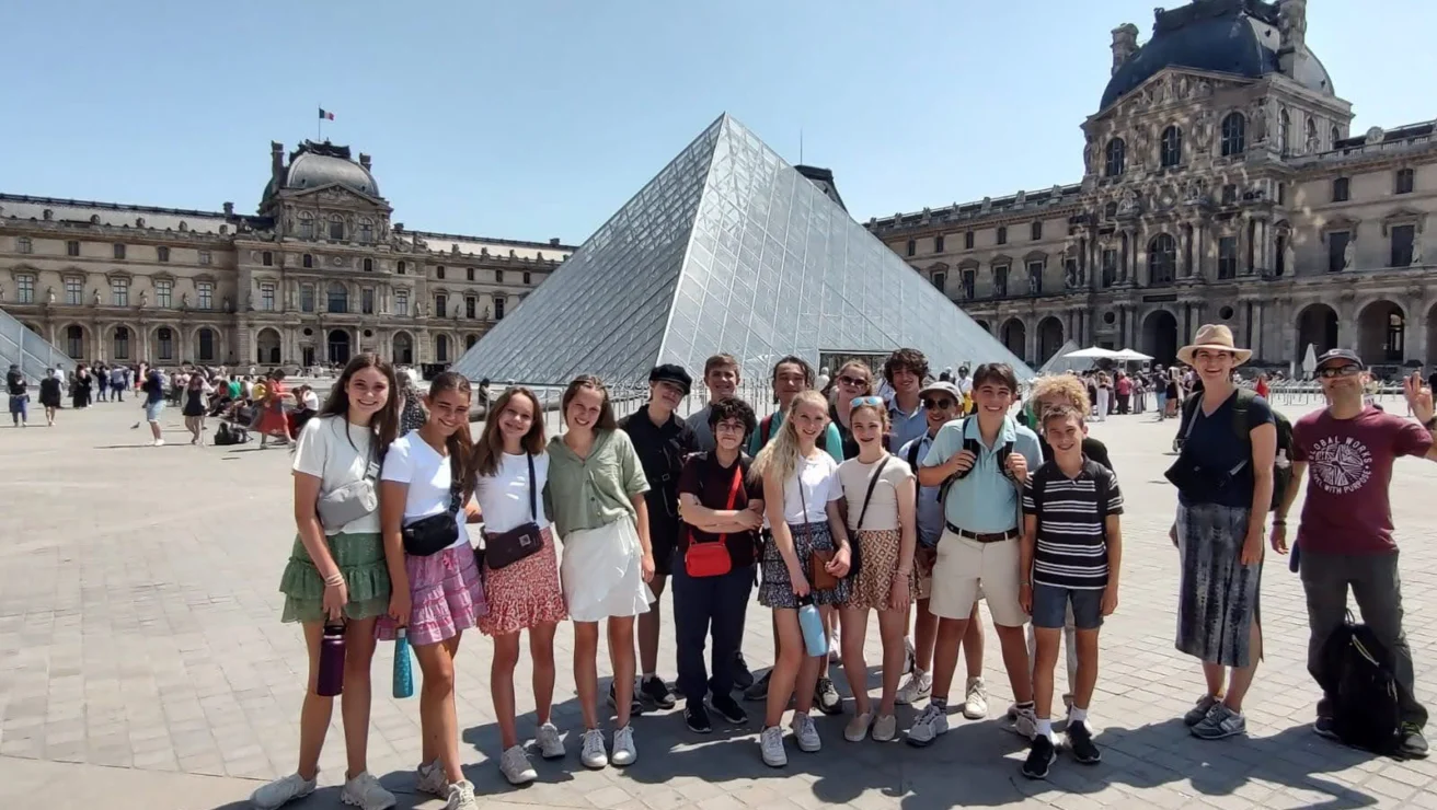 A group of people stands in front of the glass pyramid at the Louvre Museum in Paris, posing for a photo on a sunny day.