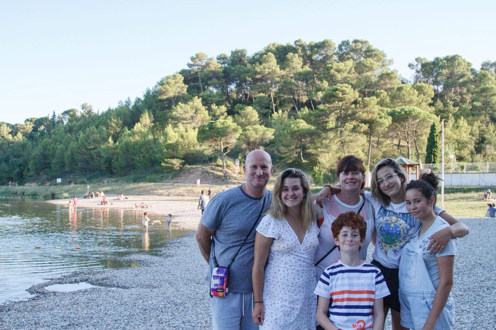 A group of six people smile on a rocky beach, with water and a forested hillside in the background.
