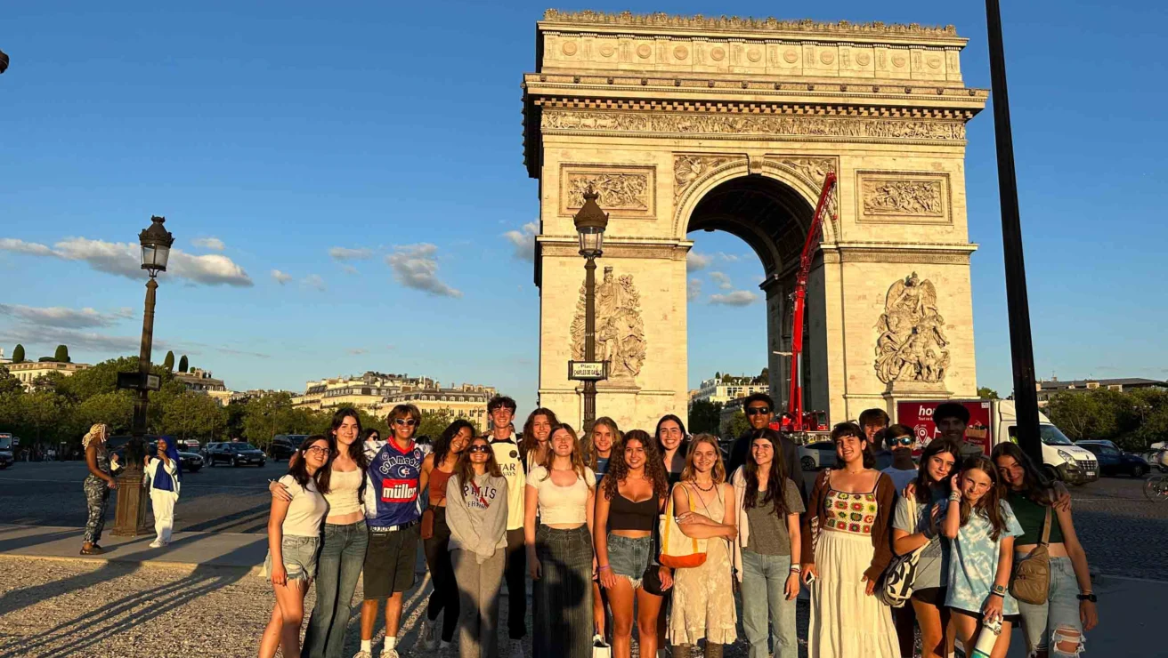 A group of people stand smiling in front of the Arc de Triomphe in Paris during a sunny day.