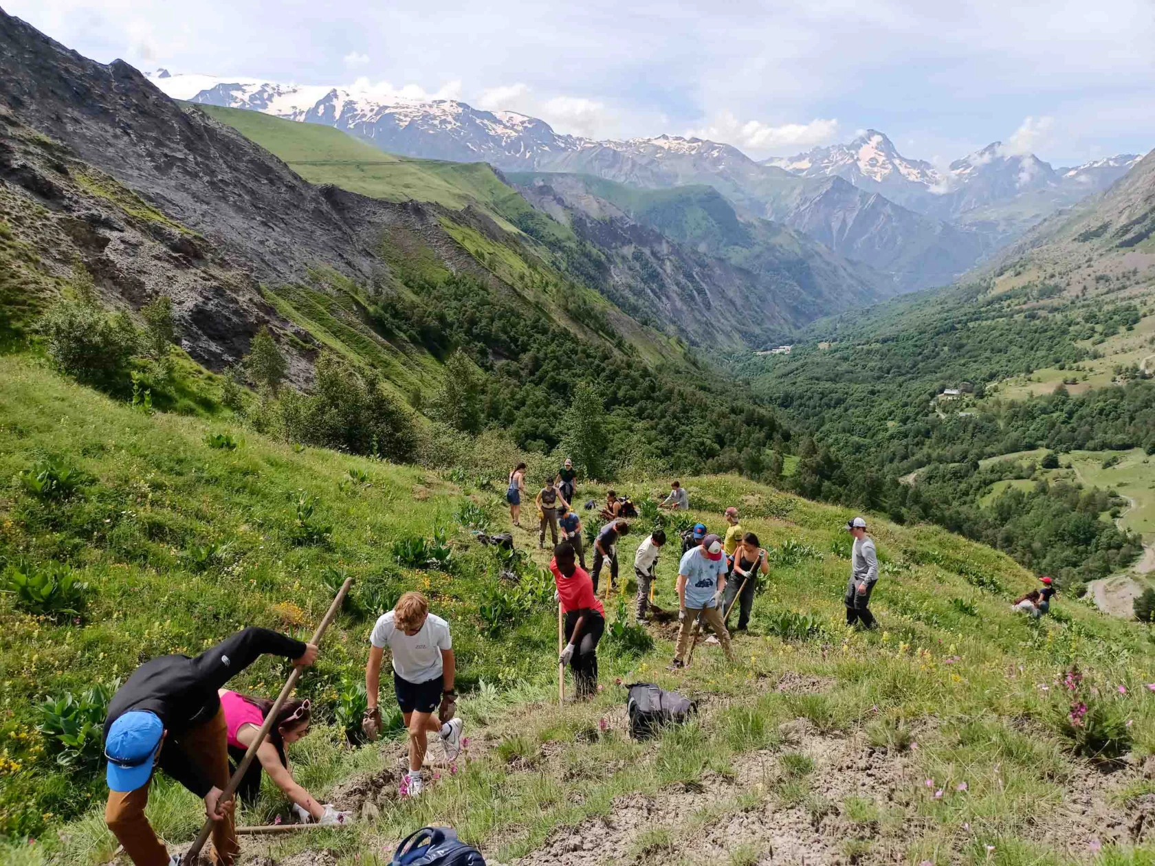 A group of people hike up a grassy mountain slope with hiking poles, surrounded by scenic views of a valley and distant snow-capped peaks under a cloudy sky.