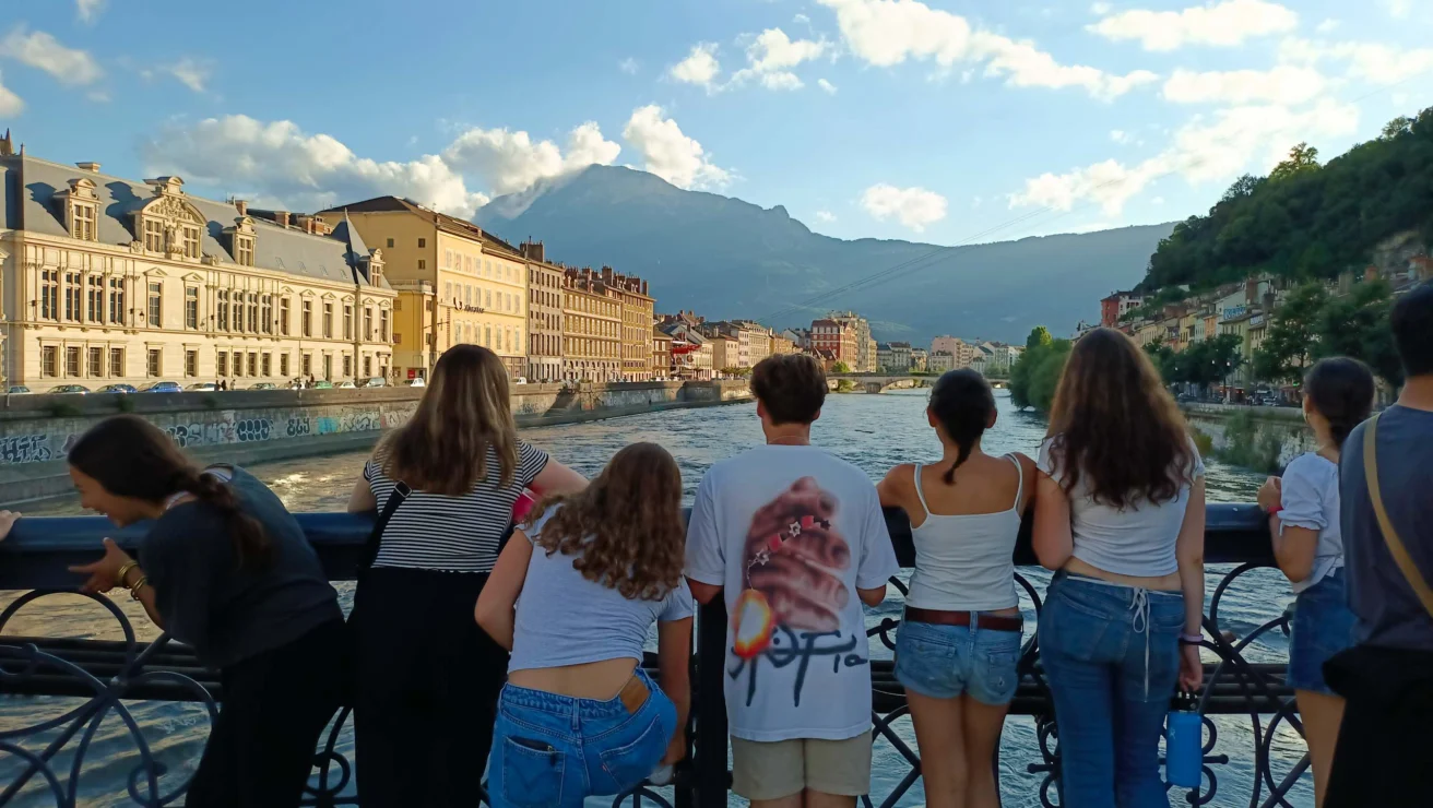 A group of people stand on a bridge railing, looking out over a river, with mountains and buildings in the background under a partly cloudy sky.