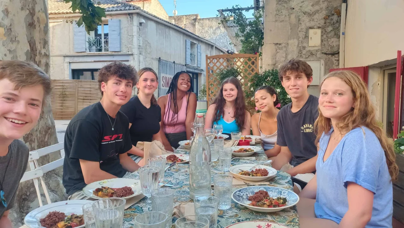 A group of eight people sits around an outdoor table, set with dishes and glasses, in a rustic setting.