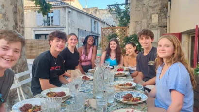 A group of eight people sits around an outdoor table, set with dishes and glasses, in a rustic setting.