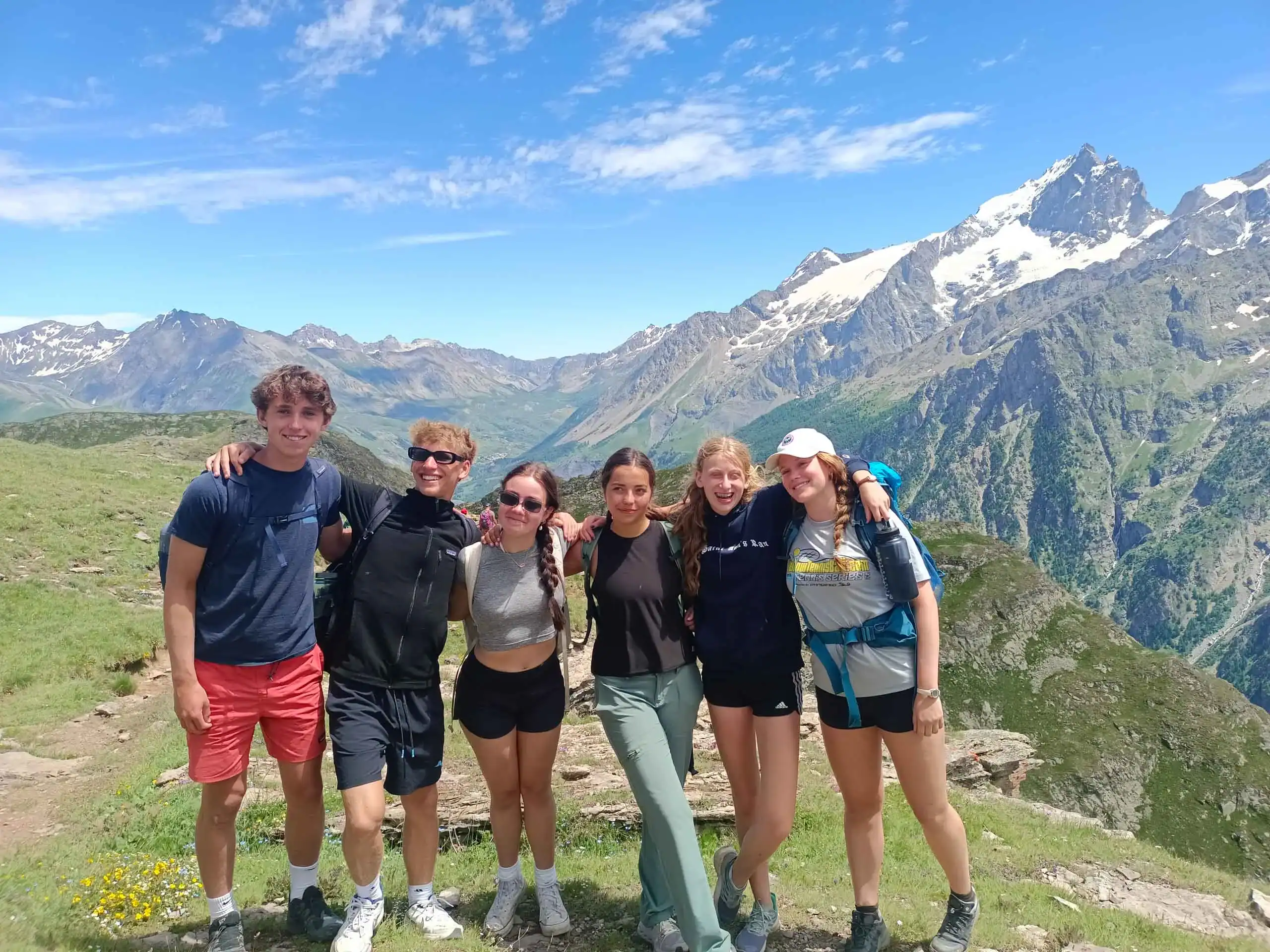 Six young students stand side by side on a grassy mountain trail, taking part in a french immersion program, with snow-capped peaks and blue sky in the background.