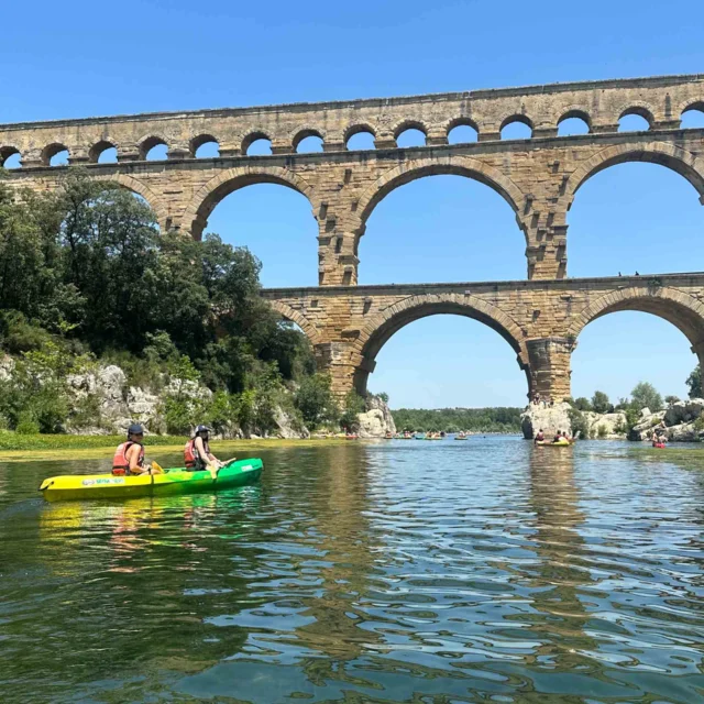 Two kayakers paddle on a river beneath an ancient stone arch bridge under a clear blue sky.