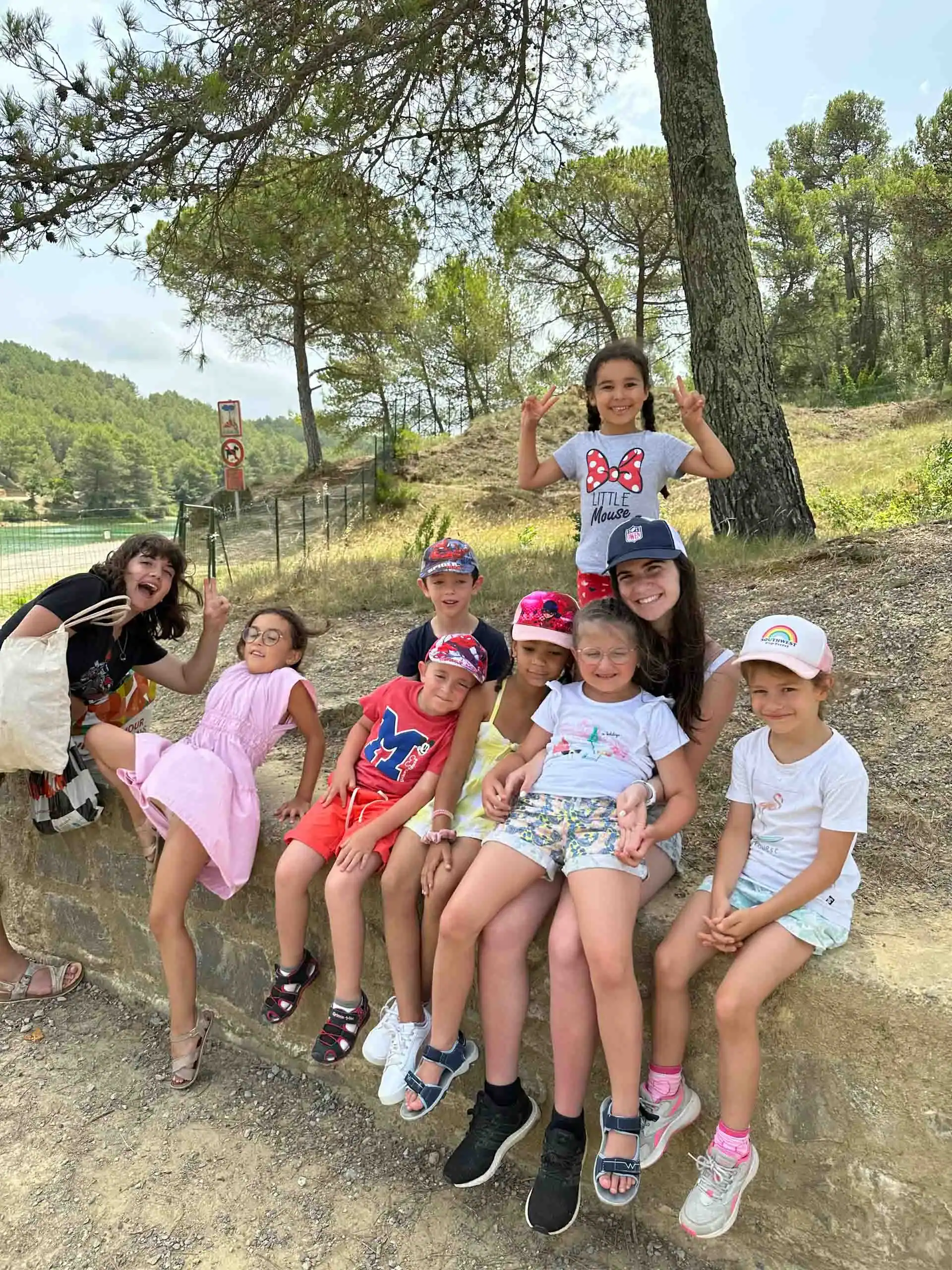 A group of children and one adult sit and pose for a photo outdoors, surrounded by trees and wearing casual summer clothes and hats during a fun French immersion program.
