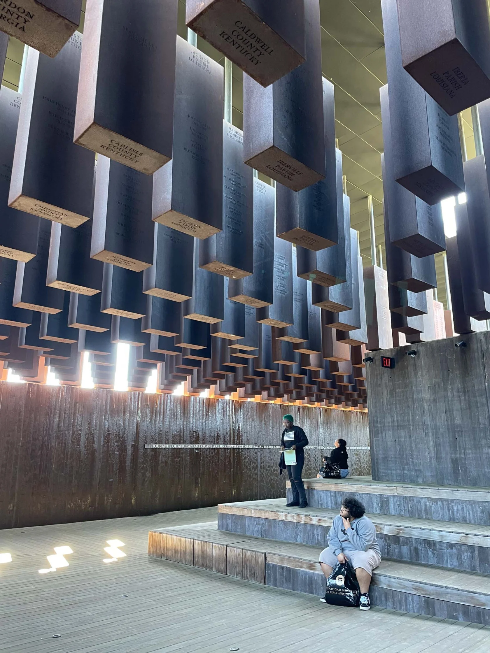 Individuals sit and stand on tiered steps under numerous suspended rectangular blocks in a sunlit, modern architectural space.