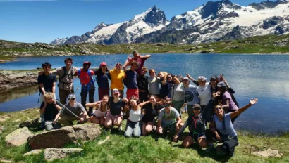 A group of people poses together on a grassy area near a lake with snow-capped mountains in the background.