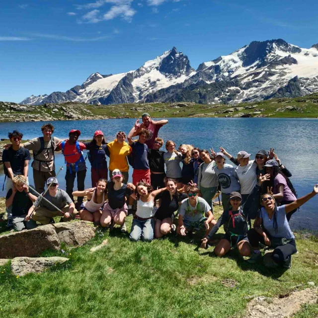 A group of people poses together on a grassy area near a lake with snow-capped mountains in the background.