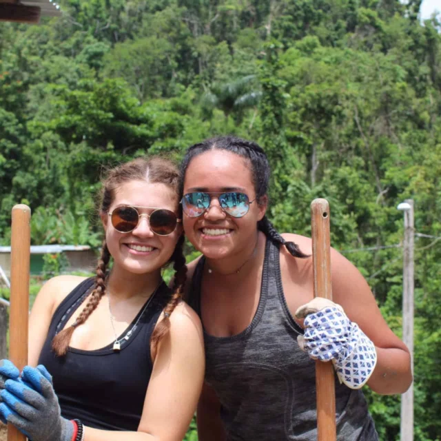 Two student travelers wearing sunglasses and gloves stand outdoors holding wooden poles, smiling at the camera with a green, forested background—enjoying a refreshing break during their school group travel adventure.