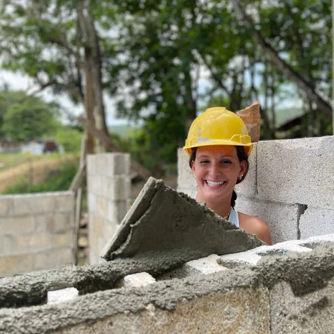 A student traveler wearing a yellow construction helmet smiles while applying mortar to a concrete block wall at a construction site, possibly as part of a school group travel experience. Trees and unfinished walls are visible in the background.