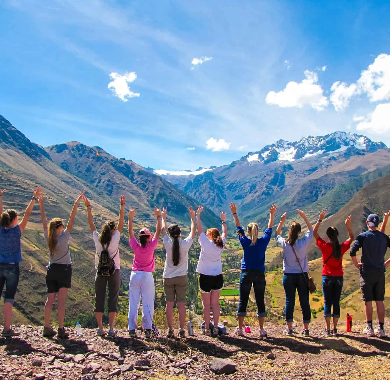 A school group travel program by Global Works brings together adventurers standing on a mountain trail, raising their arms toward snow-capped peaks and a sweeping valley beneath a clear blue sky.