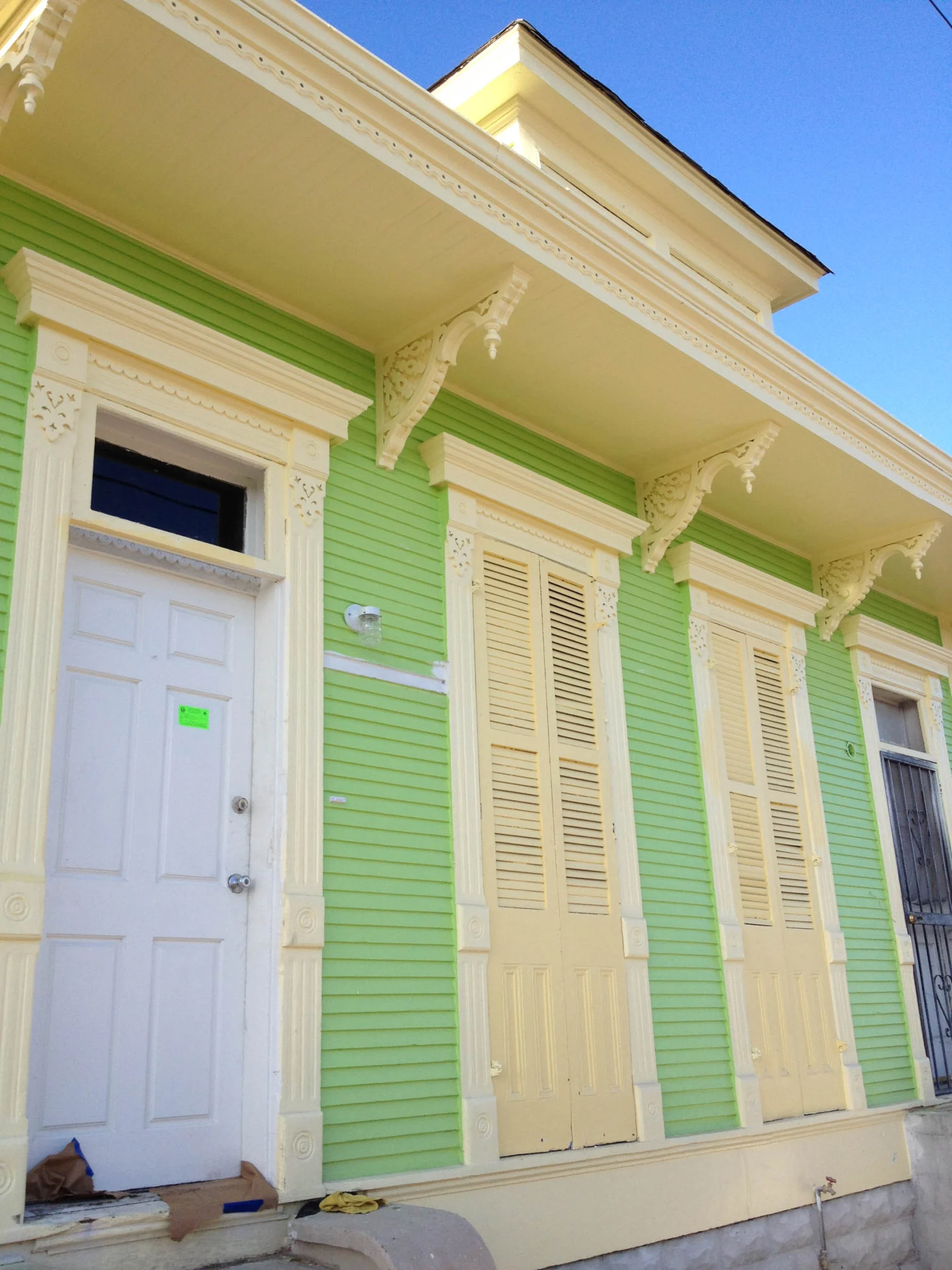 A green and yellow wooden house with decorative trim and closed shutters under a clear blue sky.