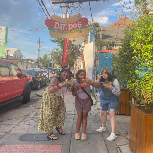 Three people enjoying hot dogs on a lively street outside a restaurant called "Dat Dog.